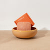 Bar of soap in a ceramic bowl with an orange packaging labeled 'Pebble' on a light wooden surface.